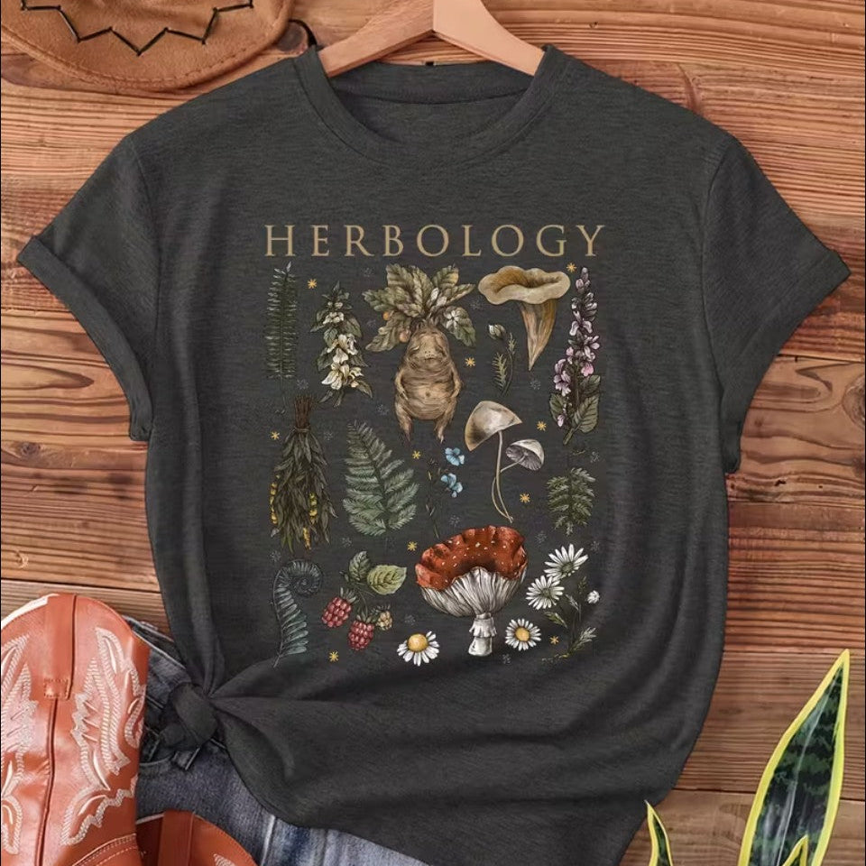 Black t-shirt with 'HERBOLOGY' design featuring plants and mushrooms on a wooden surface.