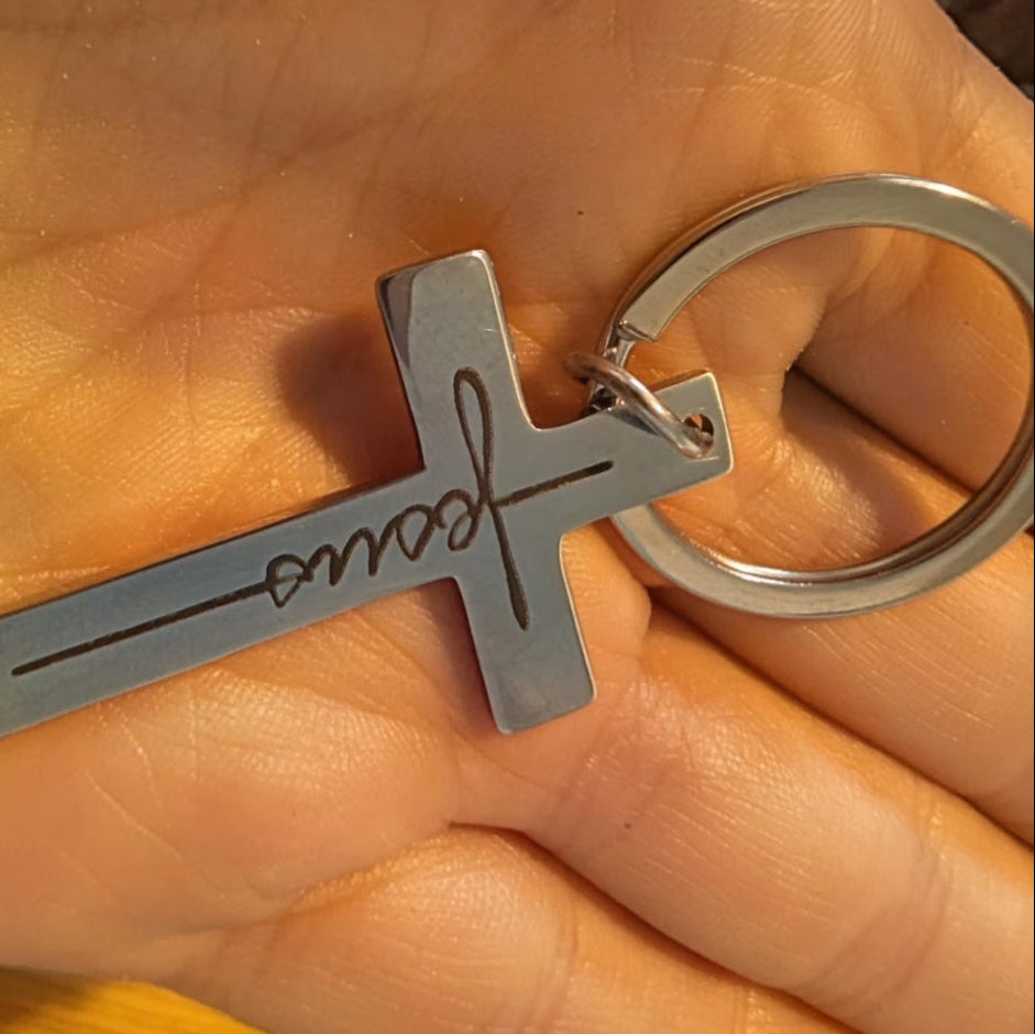 Silver cross-shaped keychain with 'Jesus' engraved on a hand.