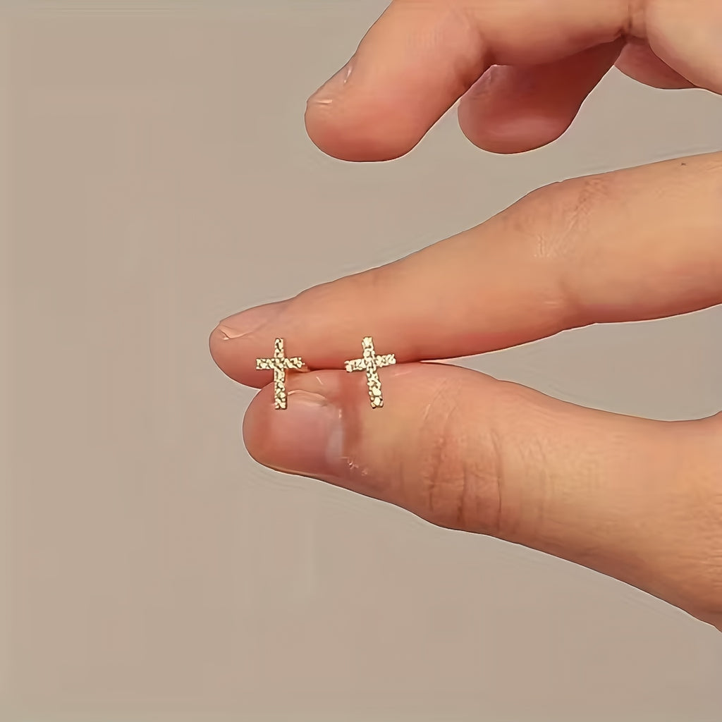 Gold cross earrings held between fingers against a neutral background
