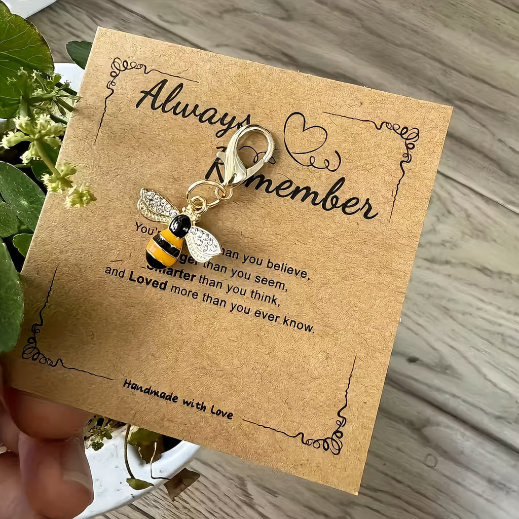 Bee-shaped keychain on a brown card with text, held over a wooden surface with a plant. always remember encouragement gift card