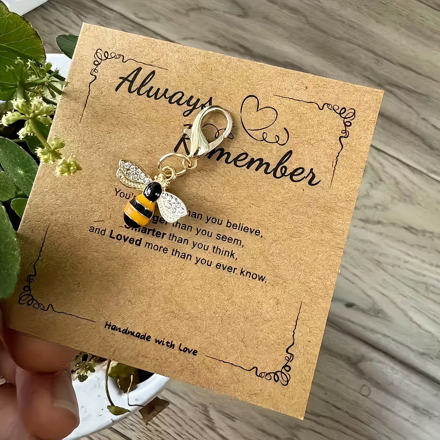 Bee-shaped keychain on a brown card with text, held over a wooden surface with a plant. always remember encouragement gift card