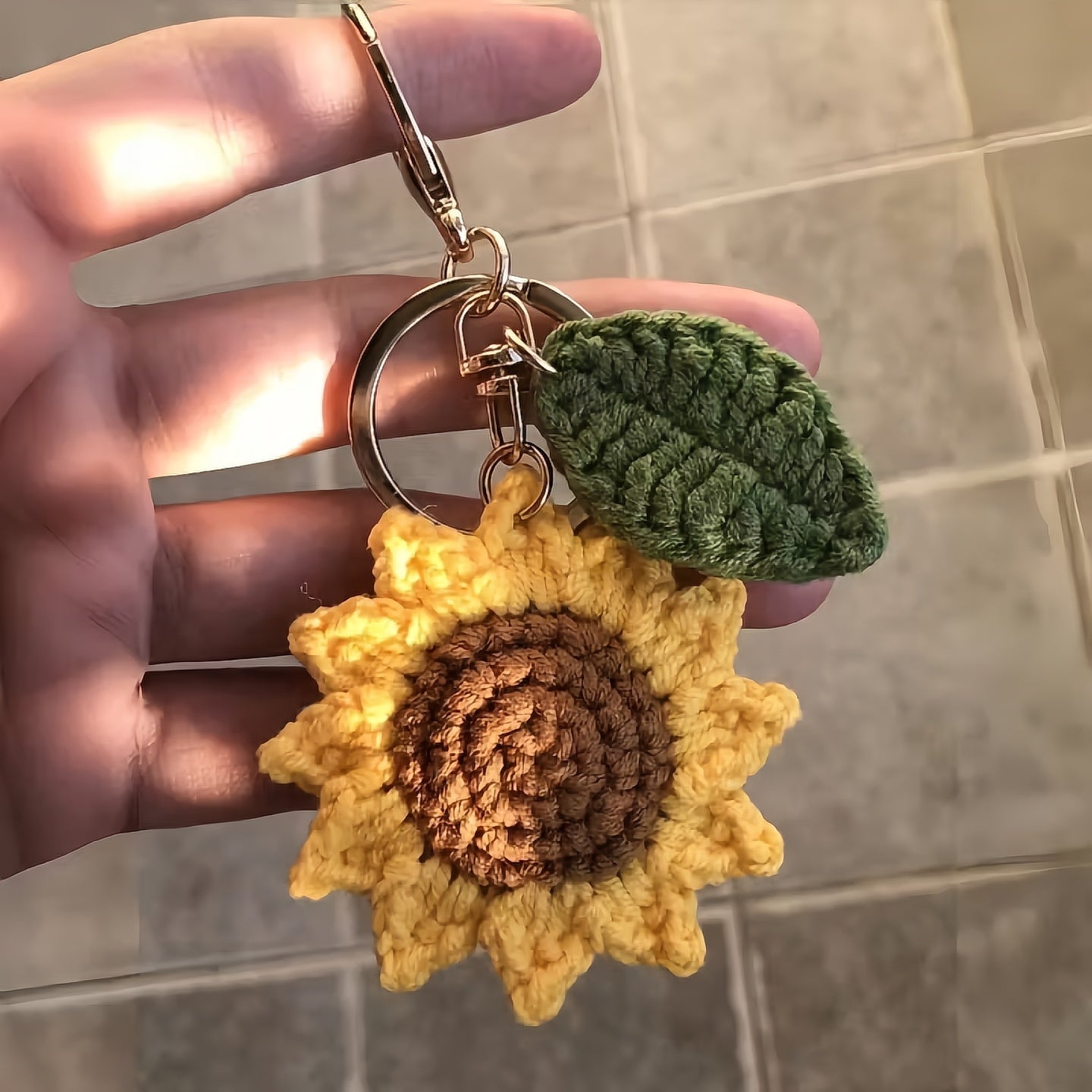 Hand holding a crocheted sunflower keychain with green leaves against a tiled wall background.