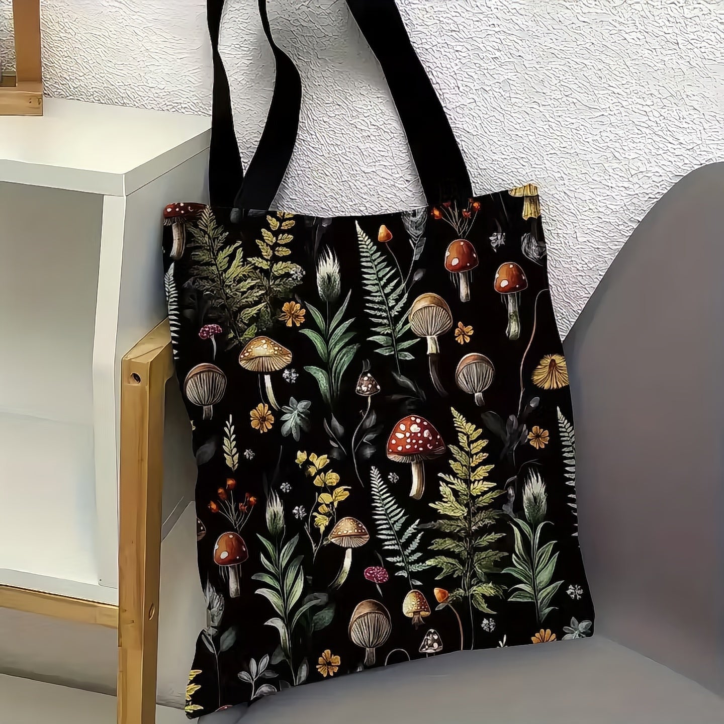 Tote bag with a forest-themed design featuring mushrooms and foliage on a black background, hanging against a white textured wall.